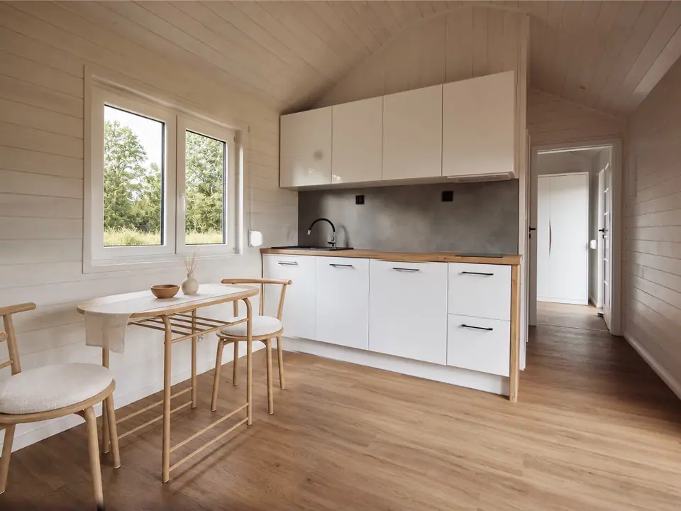 Offener Wohnbereich mit weißer Küchenzeile, davor ein ovaler Esstisch mit Holzstühlen, links ein Fenster mit Blick ins Grüne. Wände und die gewölbte Decke in heller Holzschalung. Visualisierung