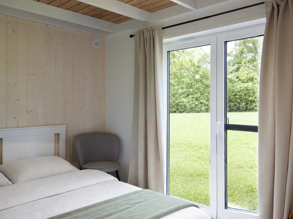 Eines der drei Schlafzimmer mit Doppelbett, weißem Holzbettgestell und grauem Polsterstuhl, einer Wandverkleidung aus hellem Fichtenholz und einer bodentiefen Terrassentür mit hellen Vorhängen, dahinter Rasen und Hecke. Visualisierung