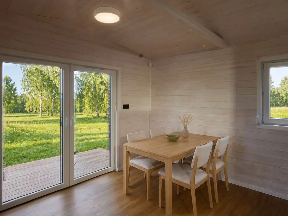 Heller Wohn- und Essbereich mit Esstisch aus Eiche und Polsterstühlen, dahinter eine bodentiefe zweiflügelige Terrassentür mit Blick auf eine Wiese mit Birken. Wände und Decke in heller Holzschalung. Visualisierung