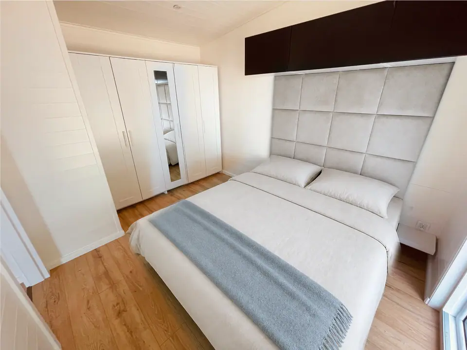 Eines der beiden Schlafzimmer mit Doppelbett, hellem Polsterkopfteil, weißem Kleiderschrank mit Spiegeltür und Hängeschränken über dem Bett. Visualisierung