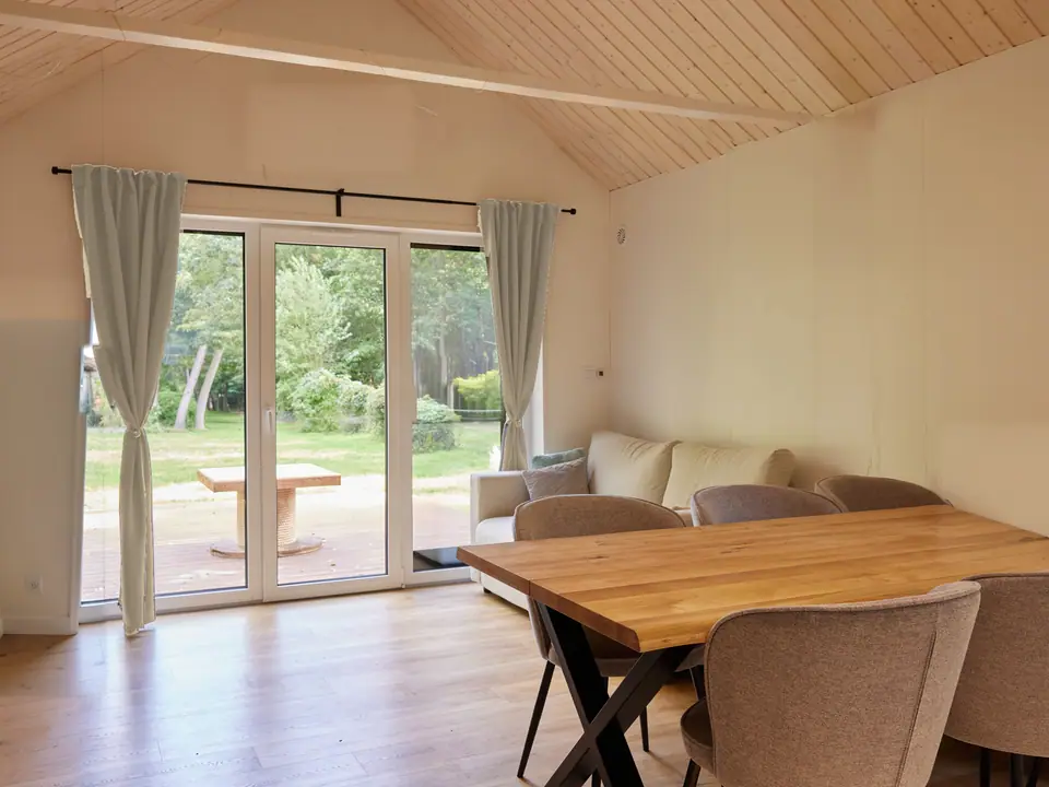 Offener Wohn- und Essbereich mit Esstisch, Polsterstühlen und hellem Sofa. Bodentiefe Terrassentür mit hellen Vorhängen, dahinter Holzterrasse und Wiese. Decke mit Holzschalung und weißem Balken. Visualisierung