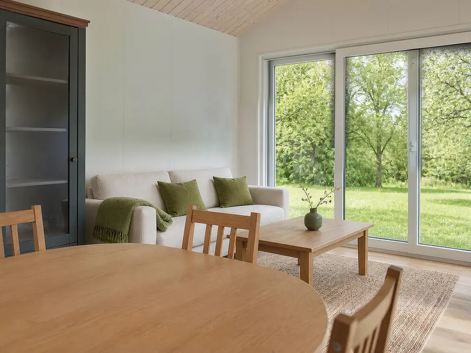Wohnbereich mit hellem Sofa, Couchtisch aus Eiche und großen Schiebefenstern zum Garten, im Vordergrund der runde Esstisch. Visualisierung